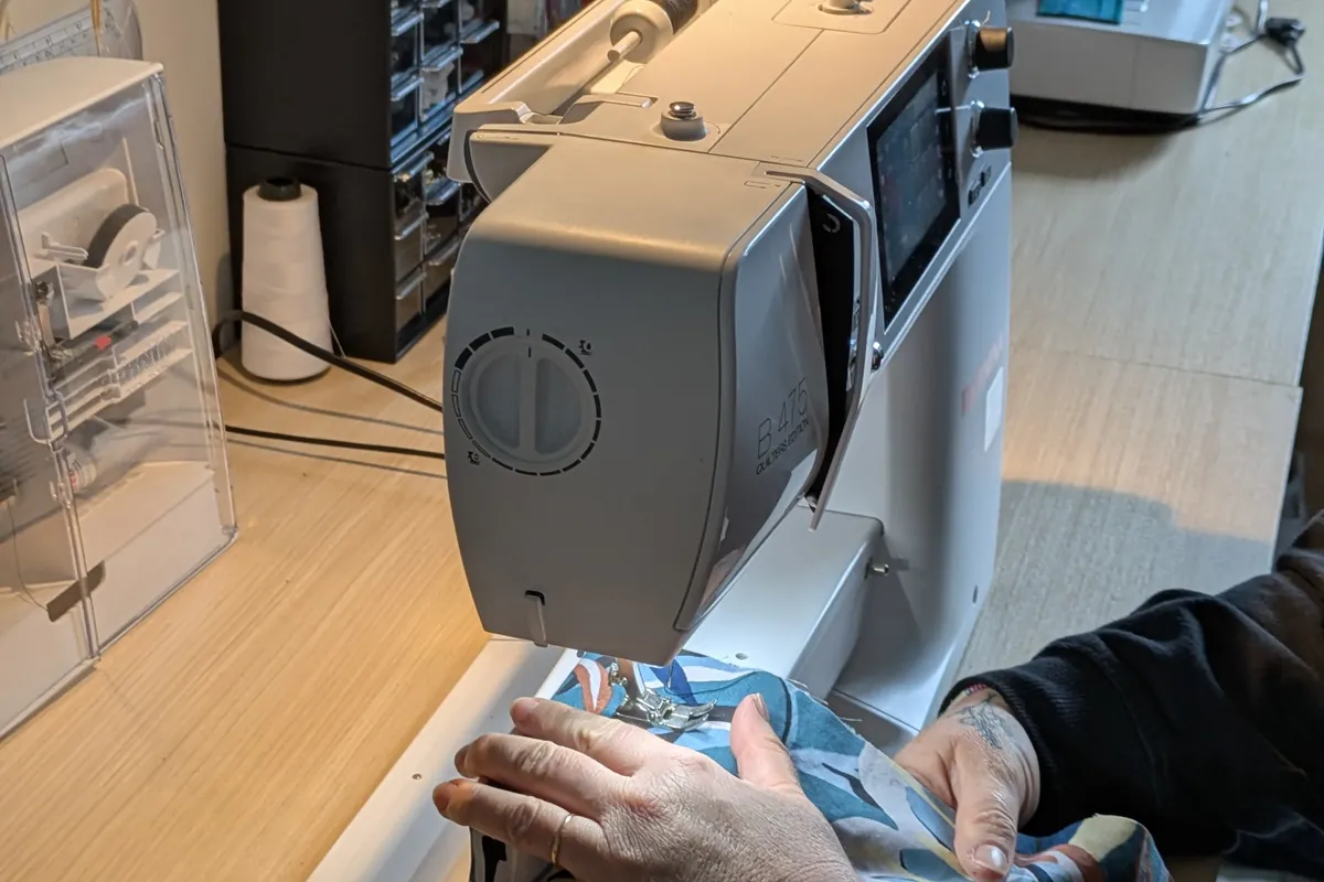 Dans un atelier lumineux, une femme coud un tissu fleuri à la machine pendant qu’une autre choisit une bobine de fil.